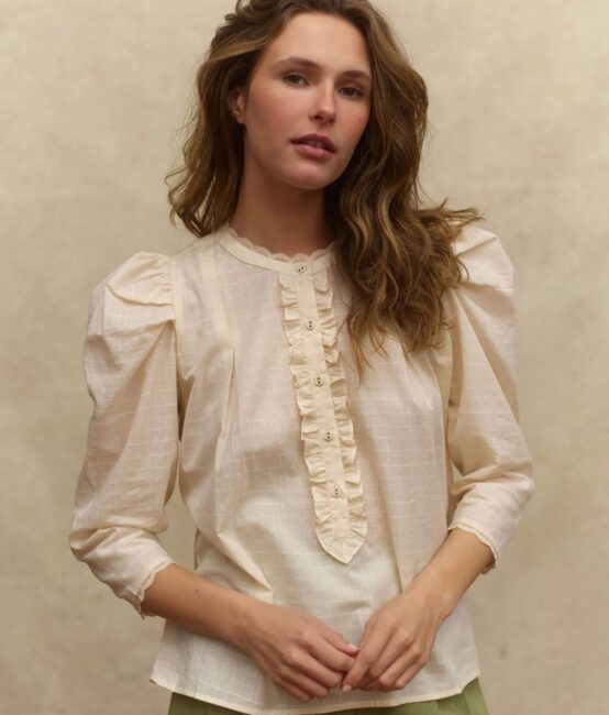 A person with long, wavy hair wears a cream-colored blouse with puffed sleeves and lace detailing, standing against a neutral background.