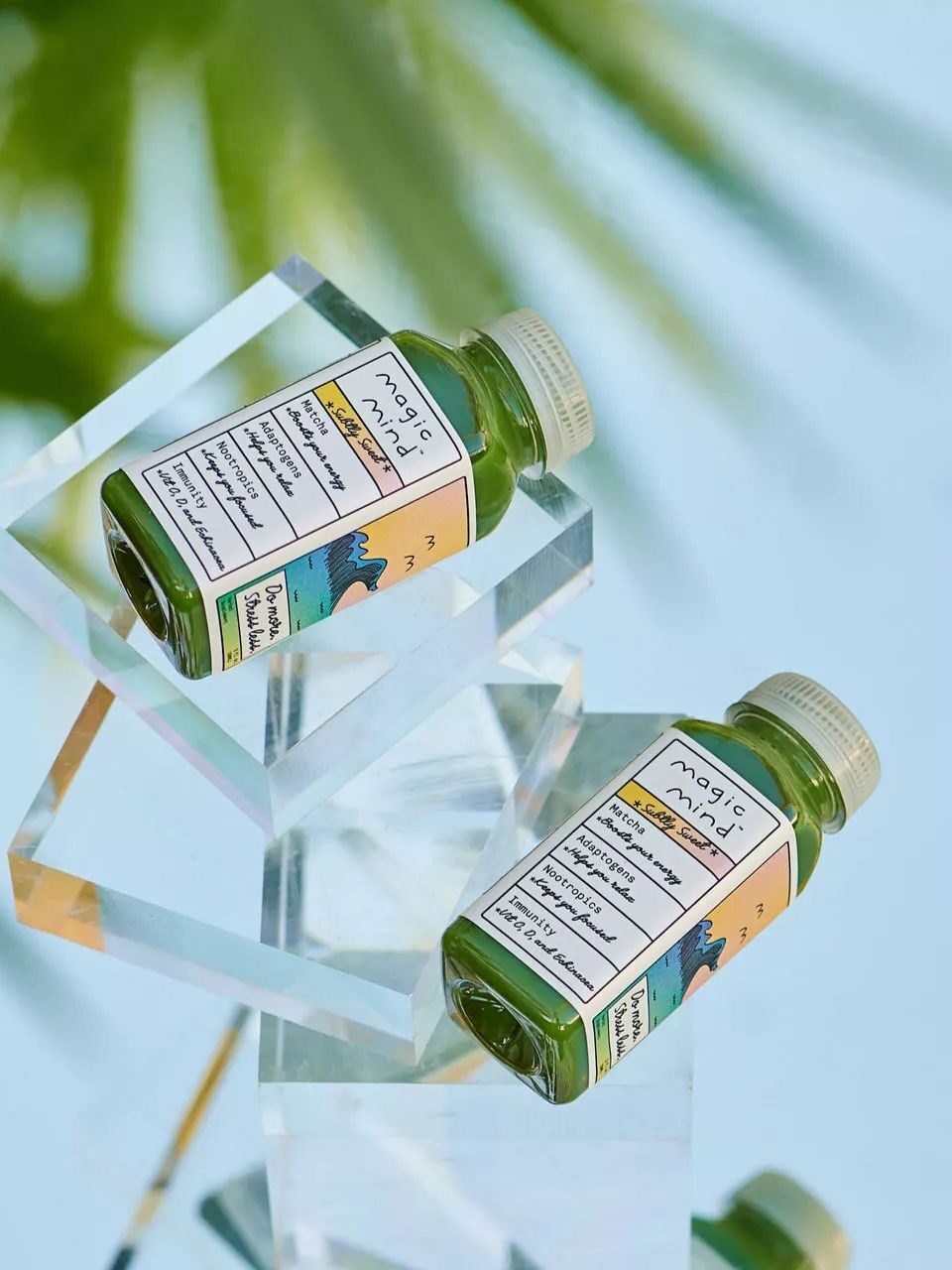 Two bottles of Magic Ami liquid soap are displayed on transparent geometric stands against a blurred background featuring green foliage. The bottles have green liquid and labels with colorful designs.