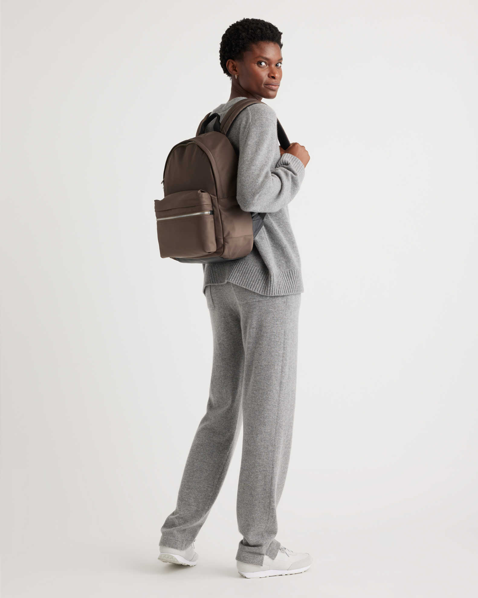 A person wearing a gray outfit and sneakers holds a brown backpack over one shoulder, standing in a relaxed pose against a plain background.