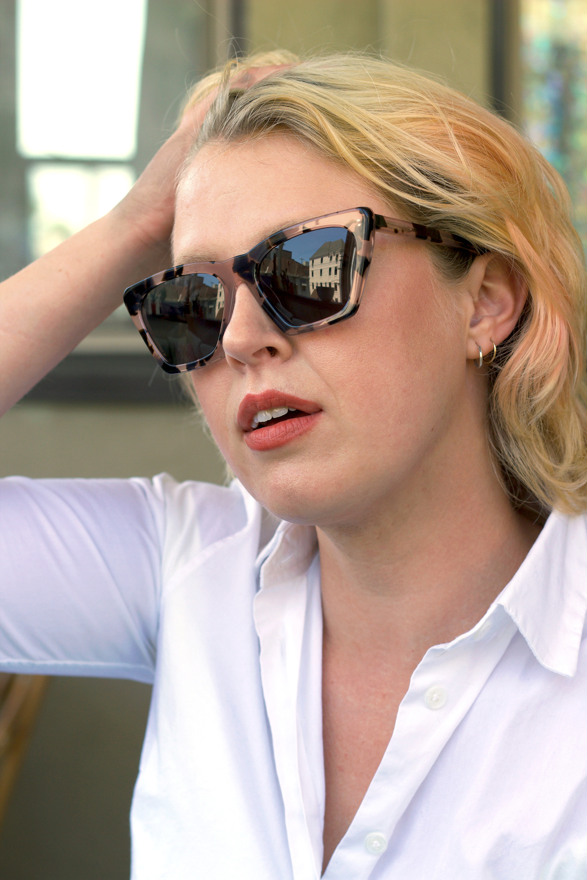 A person with blonde hair, wearing sunglasses and a white shirt, touches their head with one hand while sitting indoors.