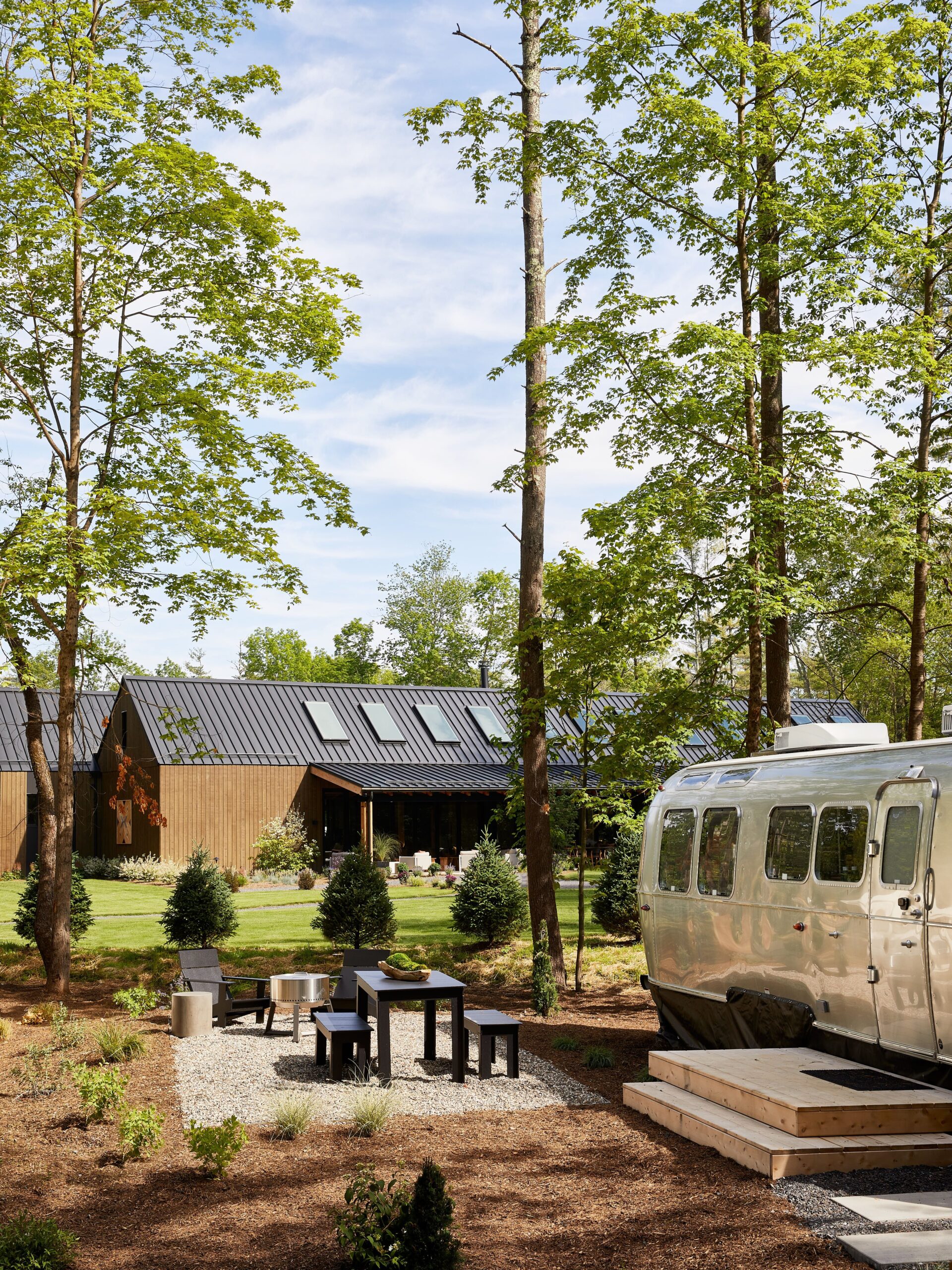 An AutoCamp Airstream set on a camping property, with a dining table and BBQ set next to it and a Clubhouse in the distance. 