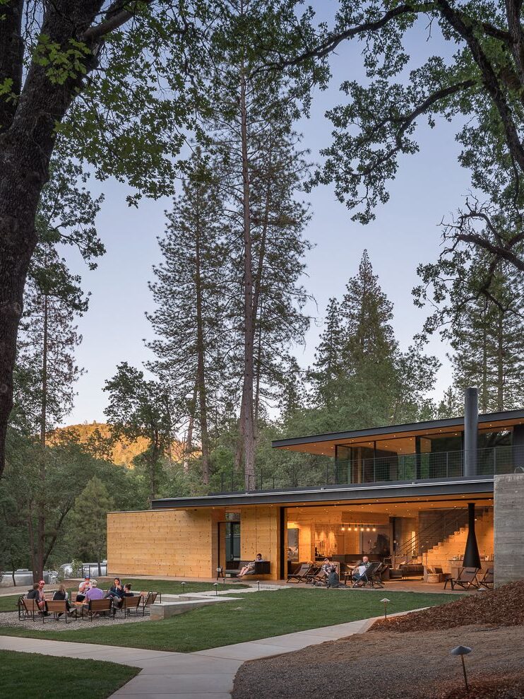 A modern house with large glass windows and wooden accents is surrounded by tall trees. Several people are seated around an outdoor fire pit on the left, and the setting sun illuminates the scene.