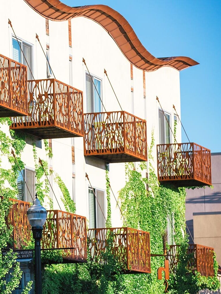 A building facade with several balconies featuring intricate metal railings, covered in green ivy against a clear blue sky.