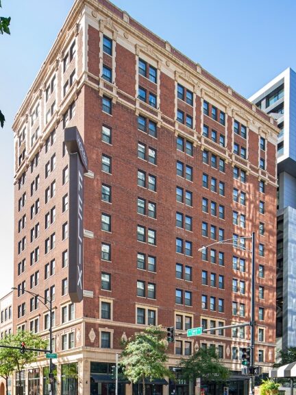 A multi-story brick building stands on a city street, surrounded by modern high-rises. Trees are present in the foreground, and a sign for "etta" is visible on the right side of the image.