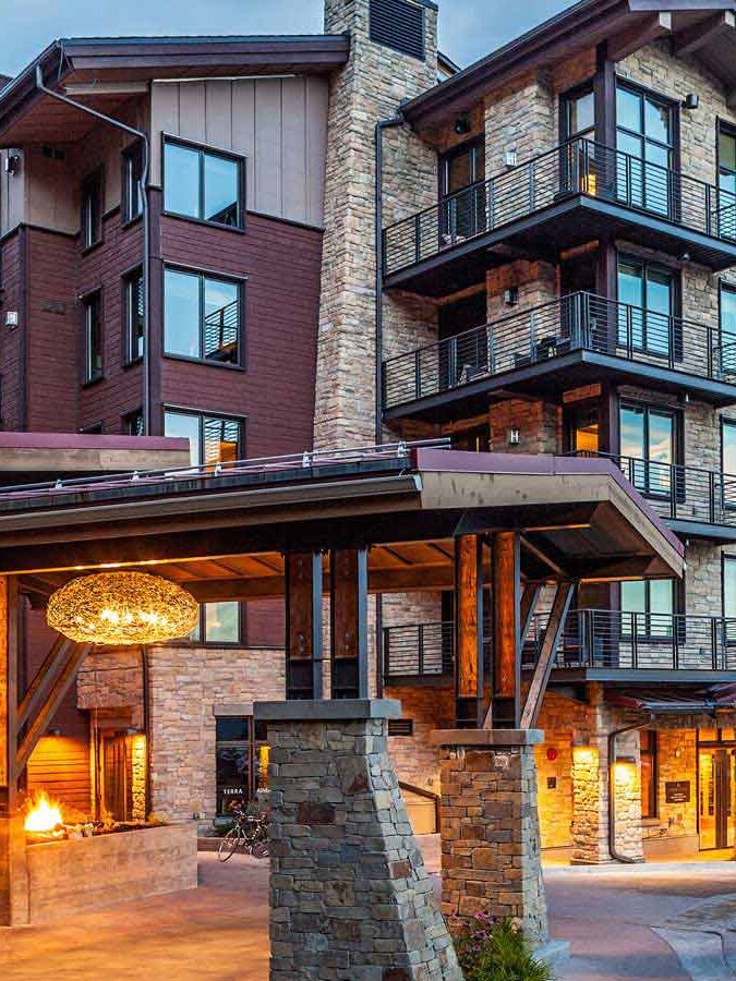 Modern multi-story hotel with large windows and balconies, featuring a stone and wood exterior. An entrance with an illuminated chandelier and a fire pit is visible in the foreground.