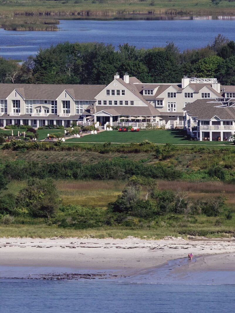 A large, multi-story seaside resort with white and beige buildings, surrounded by greenery and situated near a sandy beach with a few people walking. A body of water is visible in the background.