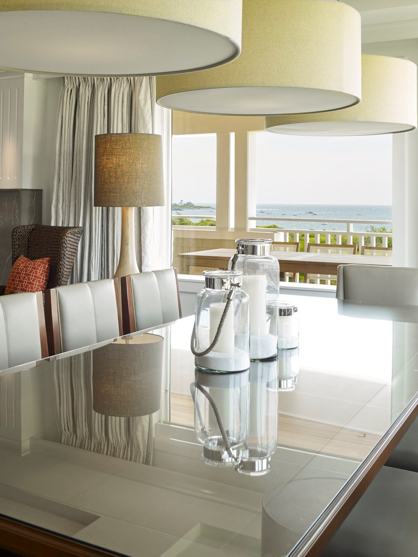 A modern dining room with a glass table, cushioned chairs, large ceiling lamps, a fireplace, and a television. Large windows offer a view of an outdoor balcony and the sea in the background.