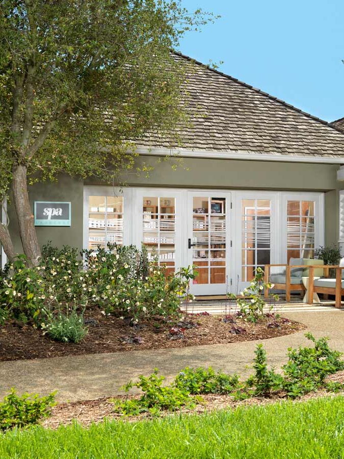 A small spa building with a wooden shingle roof and white French doors, surrounded by green shrubs and trees, with a paved walkway leading to the entrance and a seating area nearby.