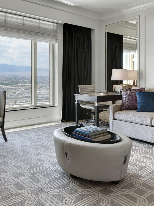 A modern hotel room with a large window offering a city view. The room features a couch, a desk, a round table with two chairs, a small oval coffee table, and wall art.