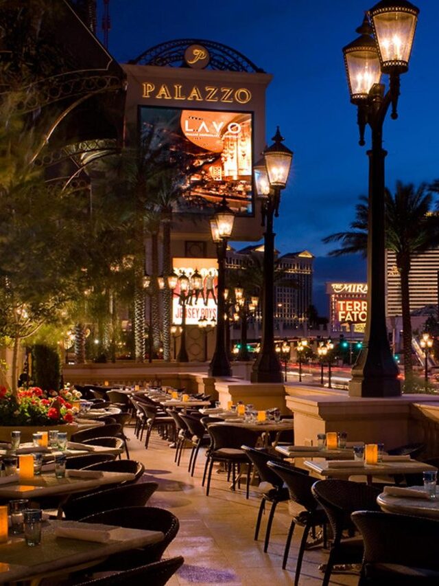 Outdoor restaurant seating area at night with lit lamps and palm trees. Background features illuminated signs for Palazzo, Treasure Island, and other Las Vegas buildings.