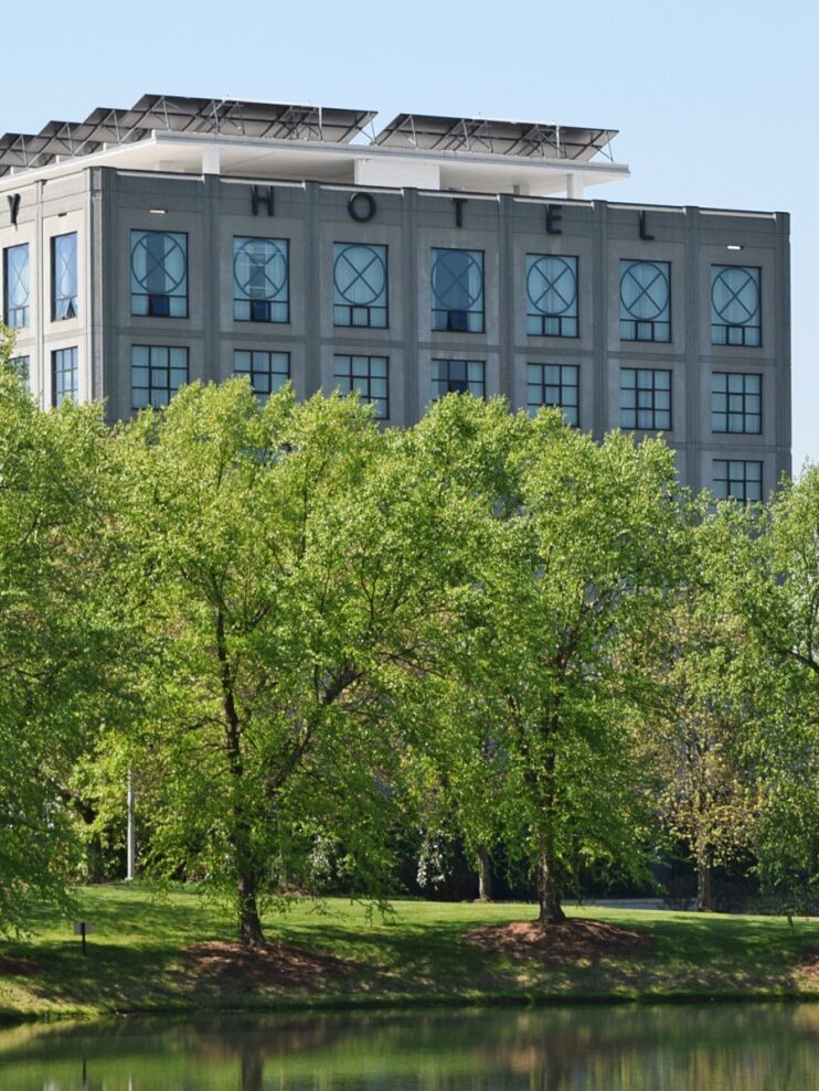 A hotel building with large windows and solar panels on the roof, partially obscured by green trees, located near a body of water on a clear day.