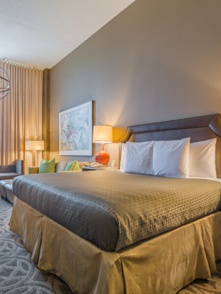 A modern hotel room with a king-sized bed, two orange bedside lamps, a small seating area, a desk with a flat-screen TV, and large window with beige curtains. The room includes abstract artwork.