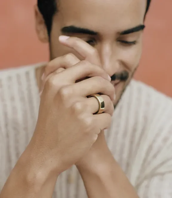 A person with a slight smile closes their eyes and clasps their hands. They are wearing a gold ring on the right hand.