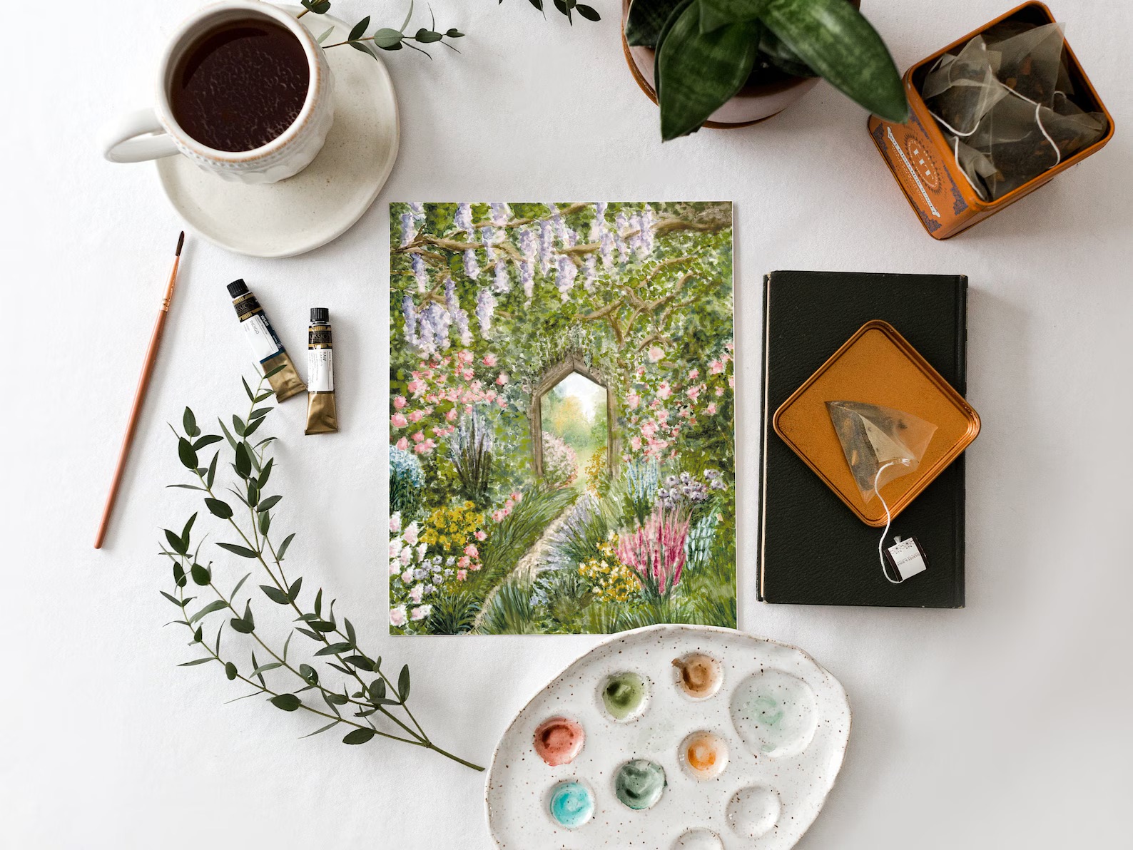 A table with a painting of a garden path, a cup of tea, tea bags, painting supplies, a palette with paint, and plants.