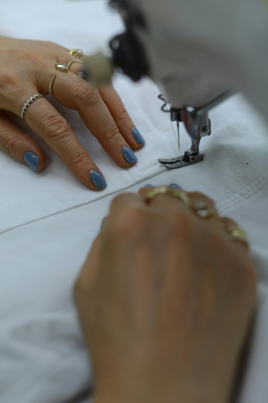 Close-up of hands wearing rings and blue nail polish, guiding white fabric under the needle of a sewing machine.