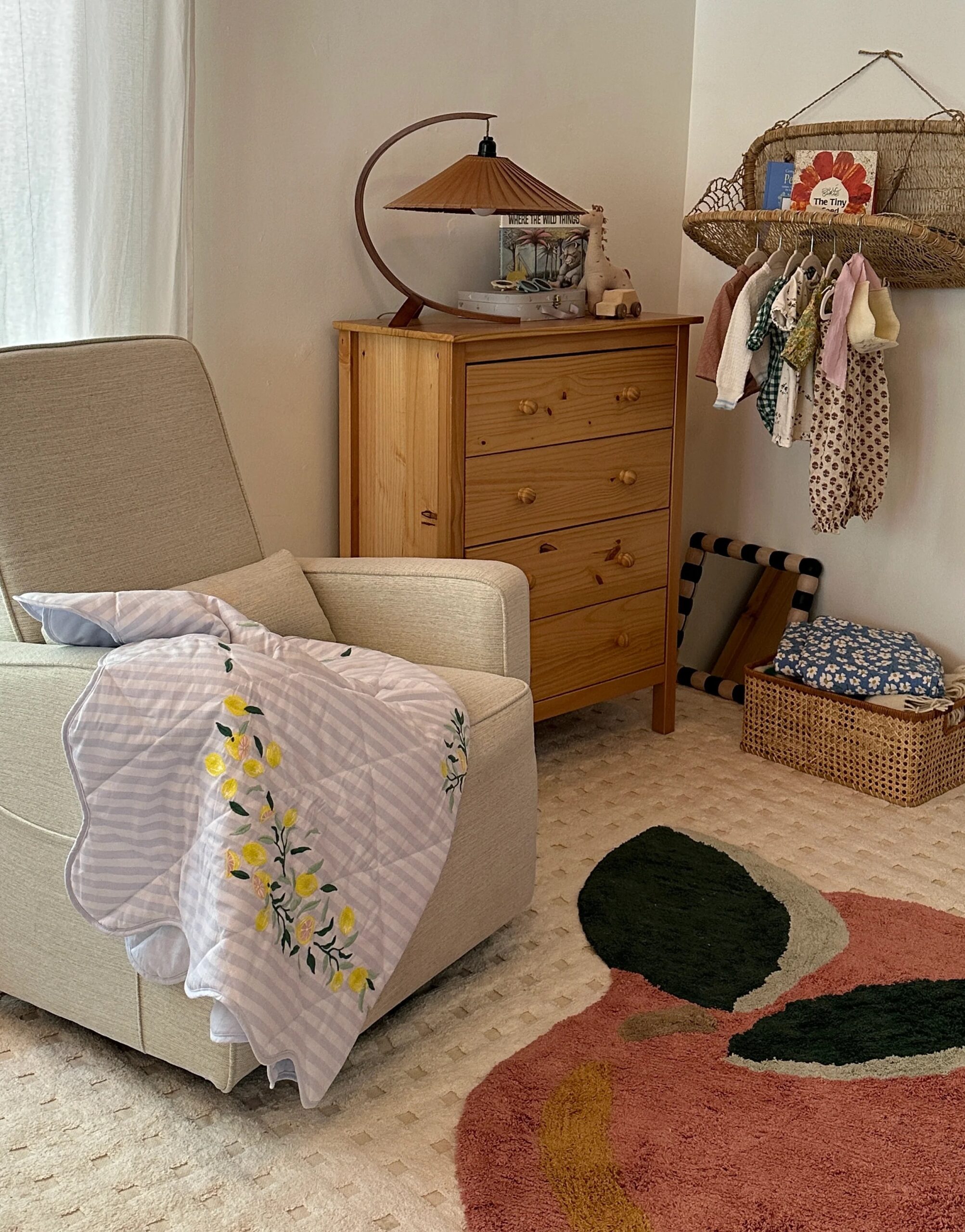 A cozy nursery with an armchair draped with a blanket, a wooden dresser with a lamp and decor, a hanging rack with baby clothes, and a patterned rug on a carpeted floor.