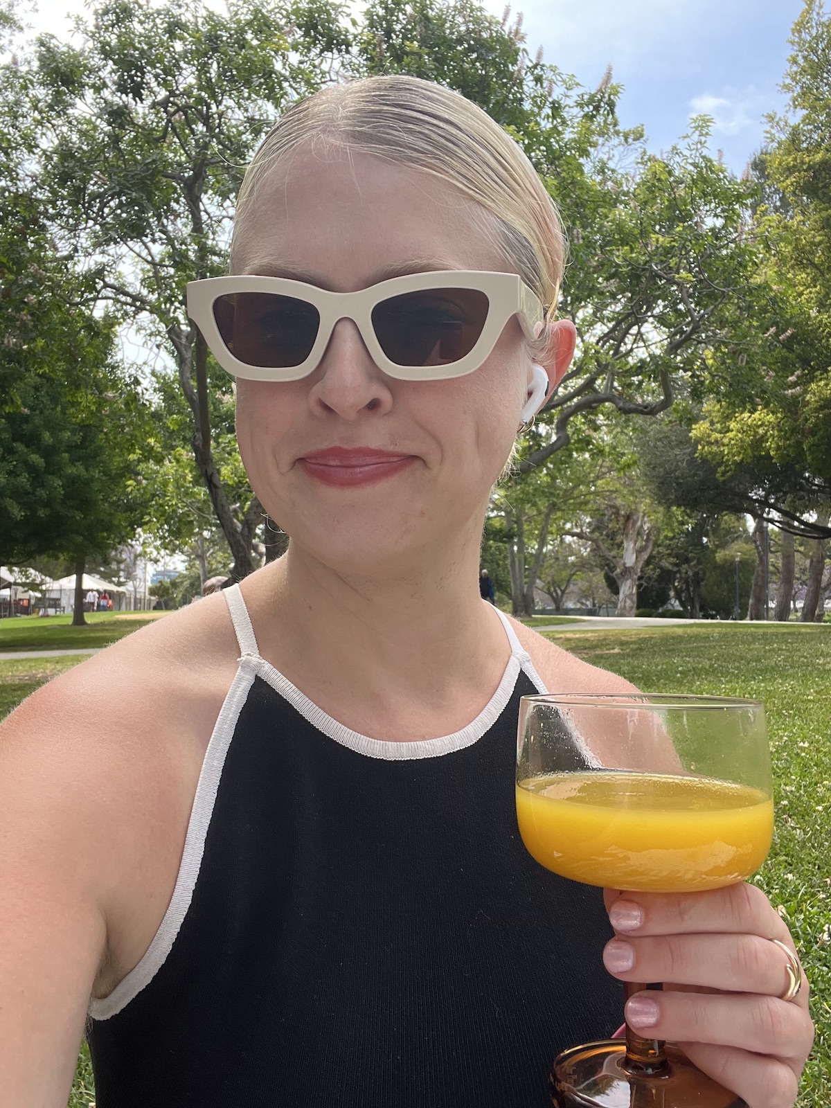 A woman wearing stylish sunglasses and a black-and-white sleeveless top enjoys her kin drink—a refreshing glass of orange juice—while sitting outdoors on a sunny day. Trees and grass are visible in the background, adding to the serene ambiance.