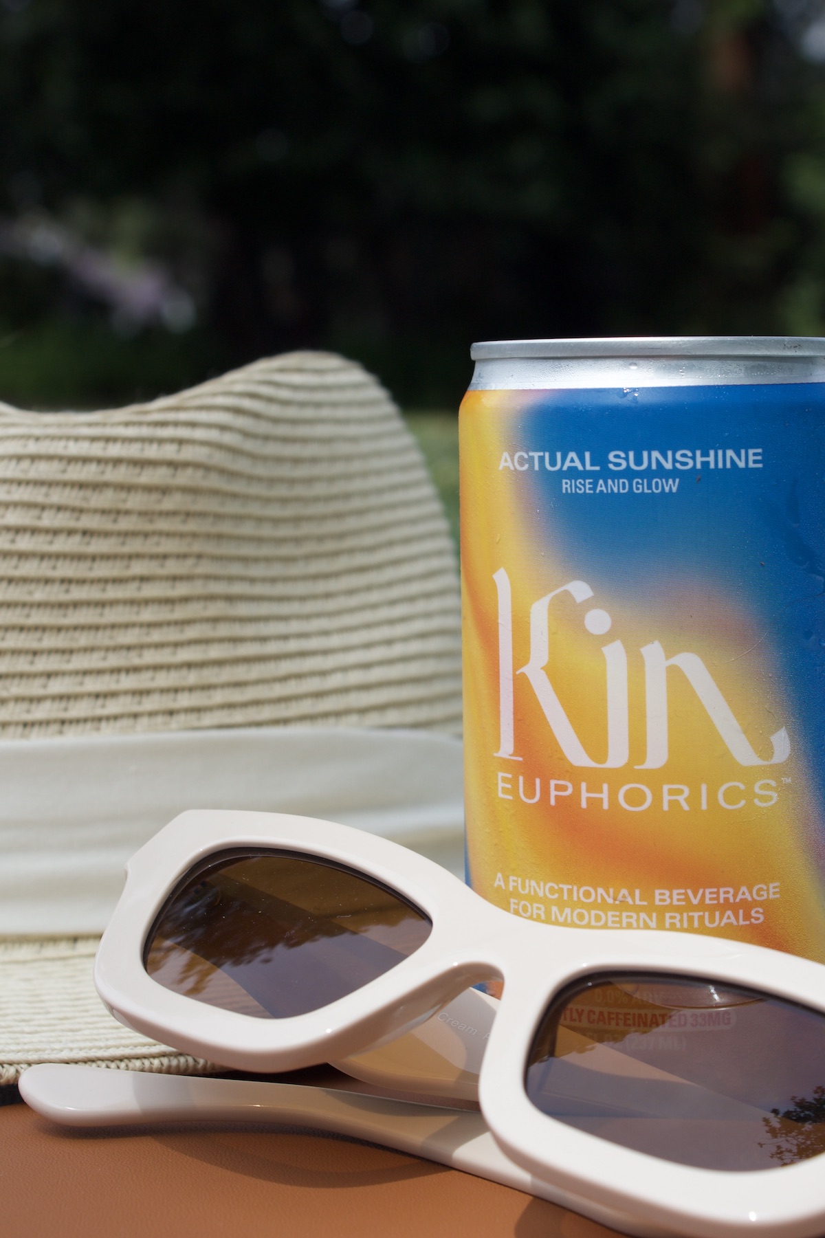 A can of Kin Euphorics beverage, labeled "Actual Sunshine," sits next to a pair of white sunglasses and a beige sun hat, showcasing the perfect summer setup with your favorite Kin drink.