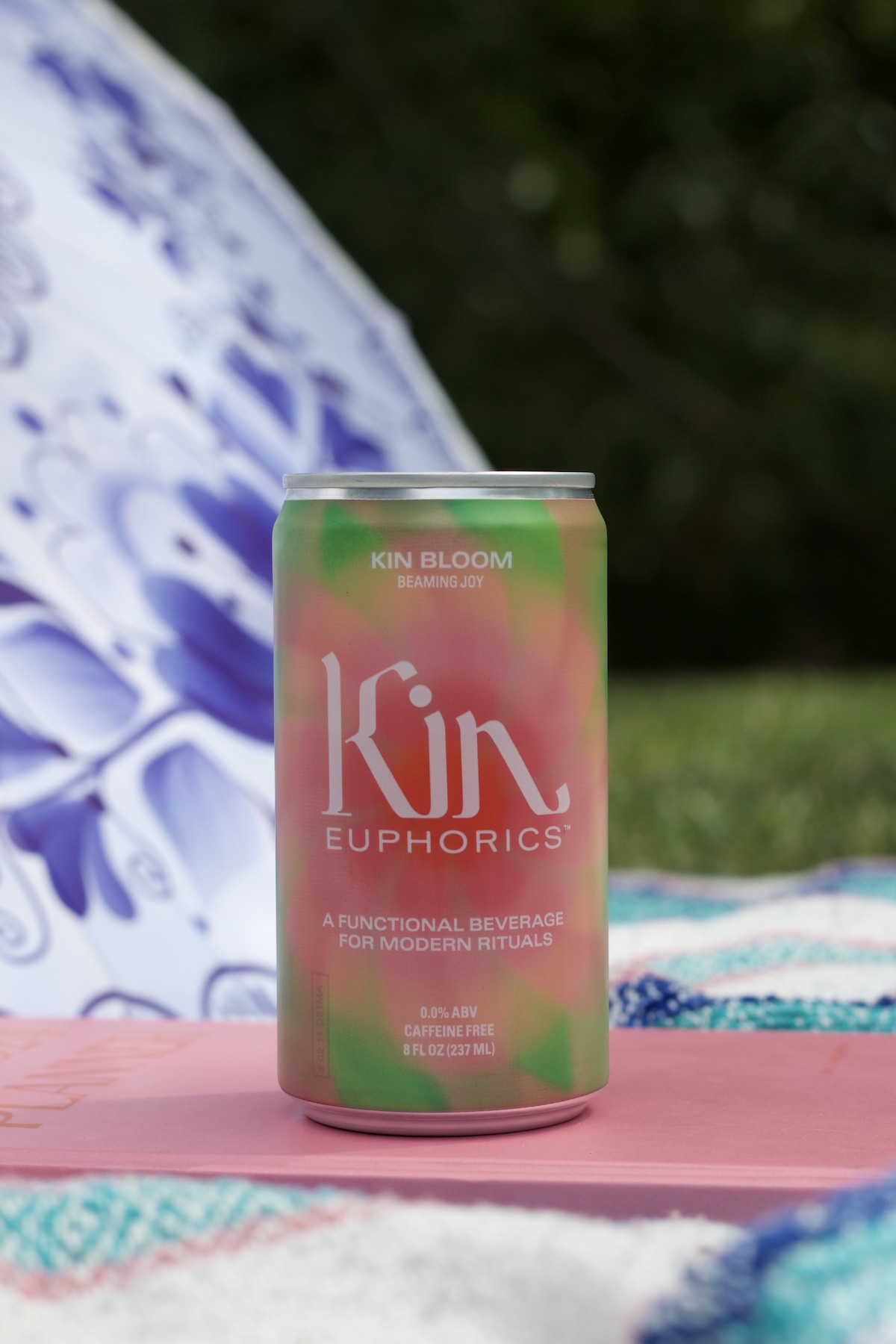 A can of Kin Euphorics' Kin Bloom, a kin drink for modern rituals, is placed on a patterned blanket outdoors against a blurred background.