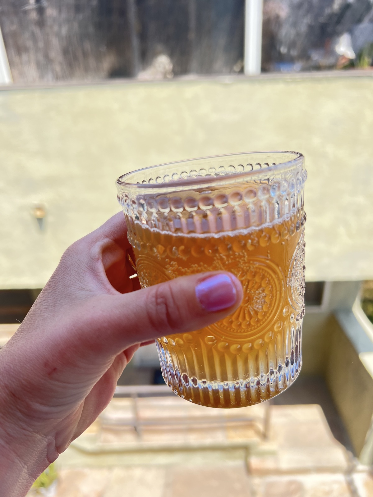 A hand with pink-painted nails holds a patterned glass of amber-colored kin drink. The background is an out-of-focus building exterior.