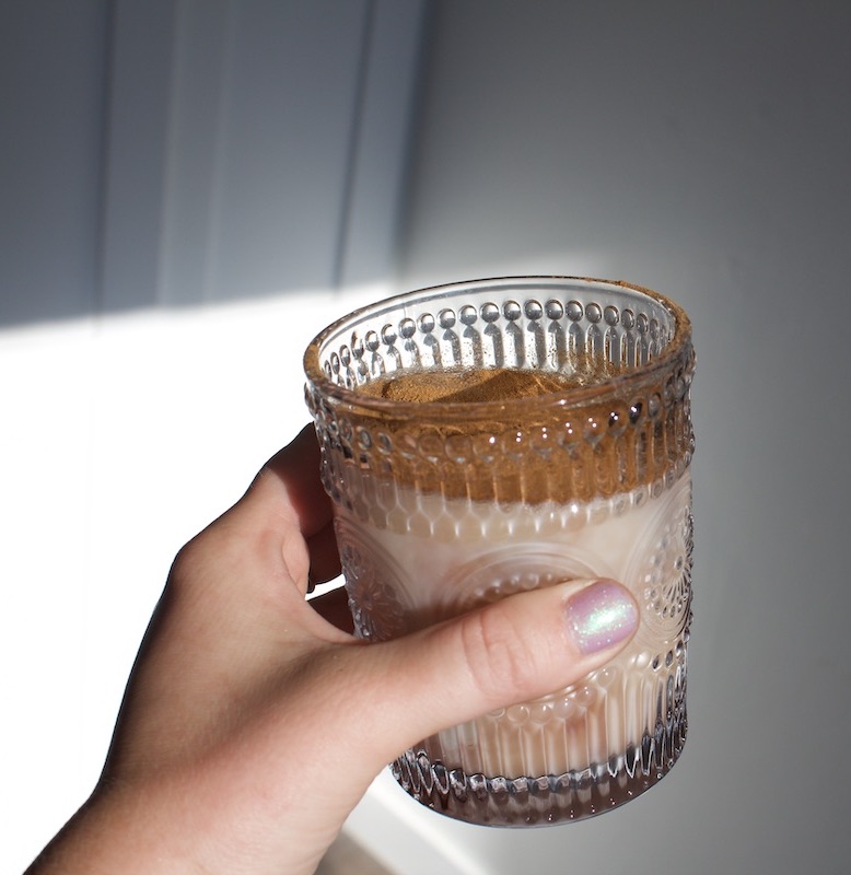 A hand holds a clear, textured glass containing a creamy Kin drink, topped with a layer of cinnamon.