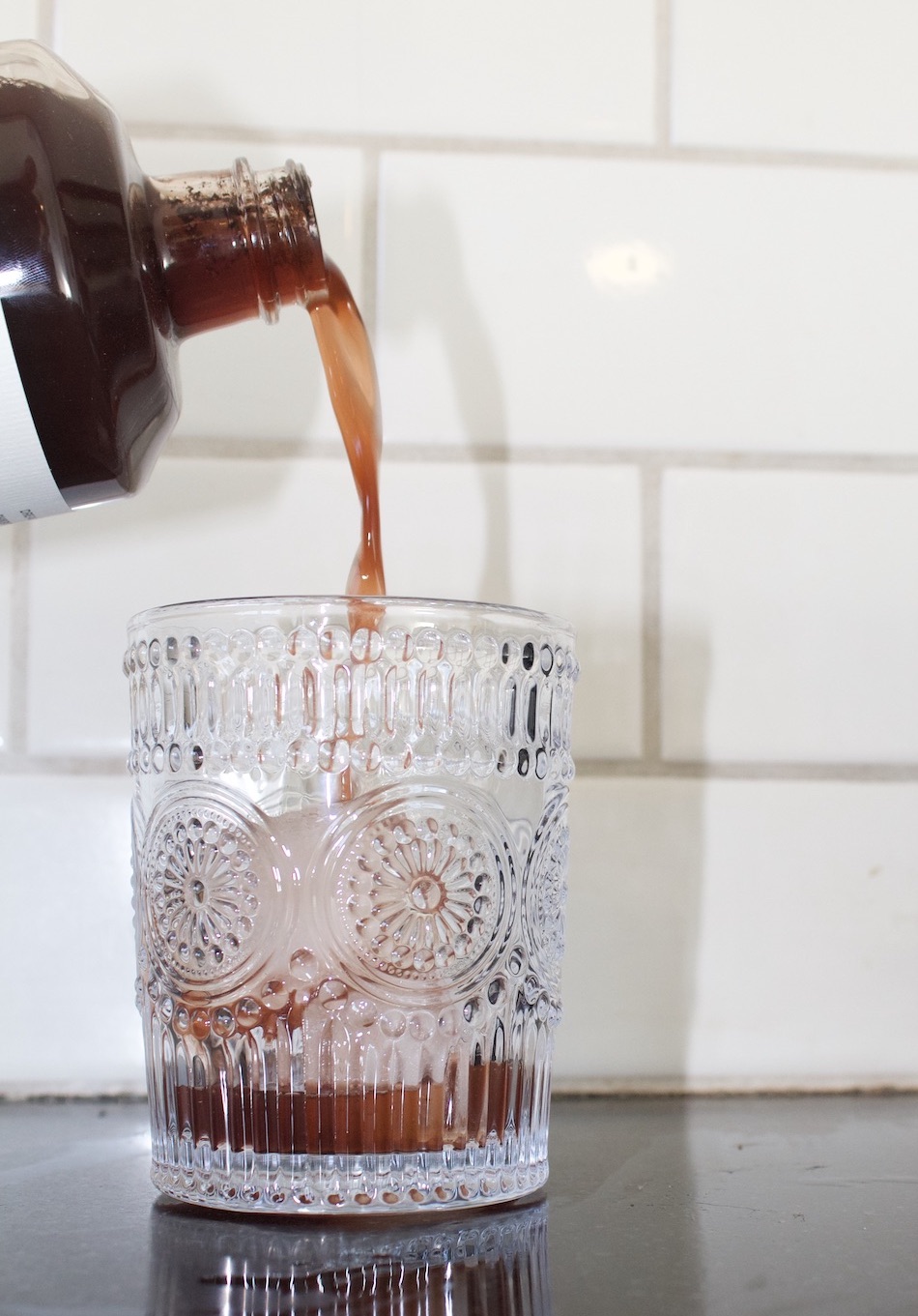 A brown liquid, perhaps a kin drink, is being poured from a bottle into a decorative glass that sits on a black surface, with a tiled wall in the background.