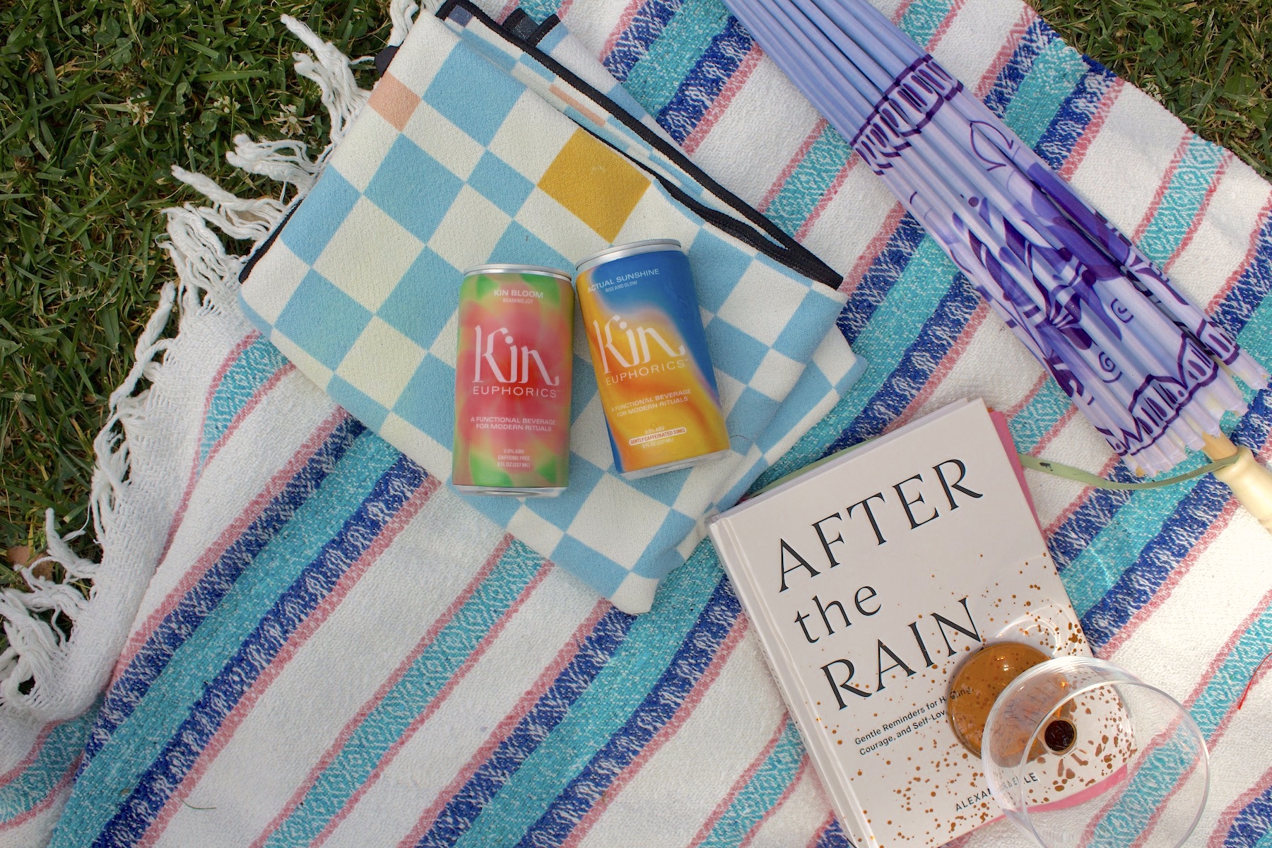 A picnic setup on a grassy lawn features a striped blanket, two Kin Euphorics cans, known as kin drinks, a book titled "After the Rain," a blue and white towel, an umbrella, and a partially filled drink glass.