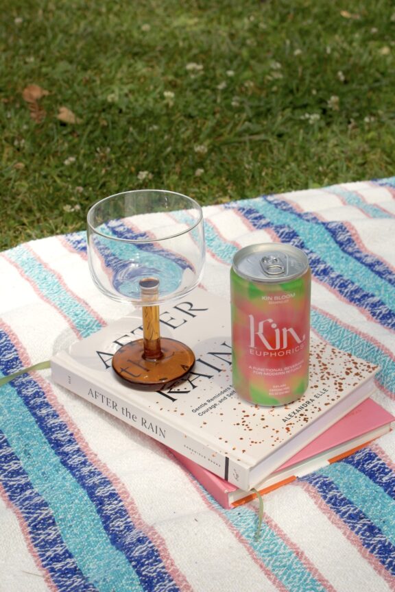 A clear glass, a can of Kin drink, and two books titled "After the Rain" are on a striped blanket on grass.