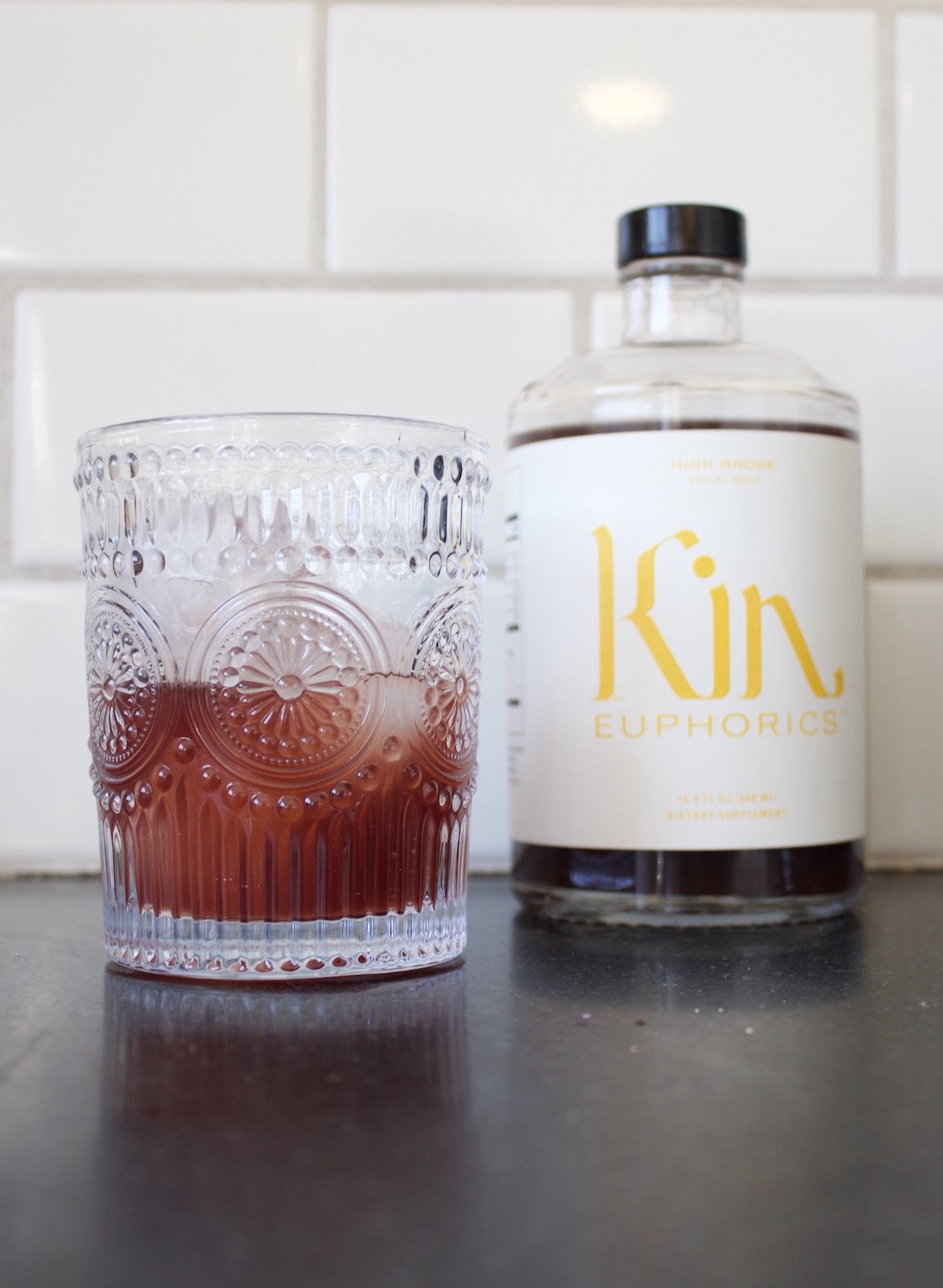 A clear glass with a textured design, partially filled with a reddish Kin drink, sits on a counter. Behind it is a bottle labeled "Kin Euphorics.