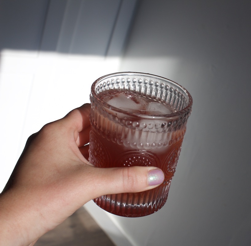 A hand holding a glass filled with a vibrant kin drink and ice cubes against a plain background.