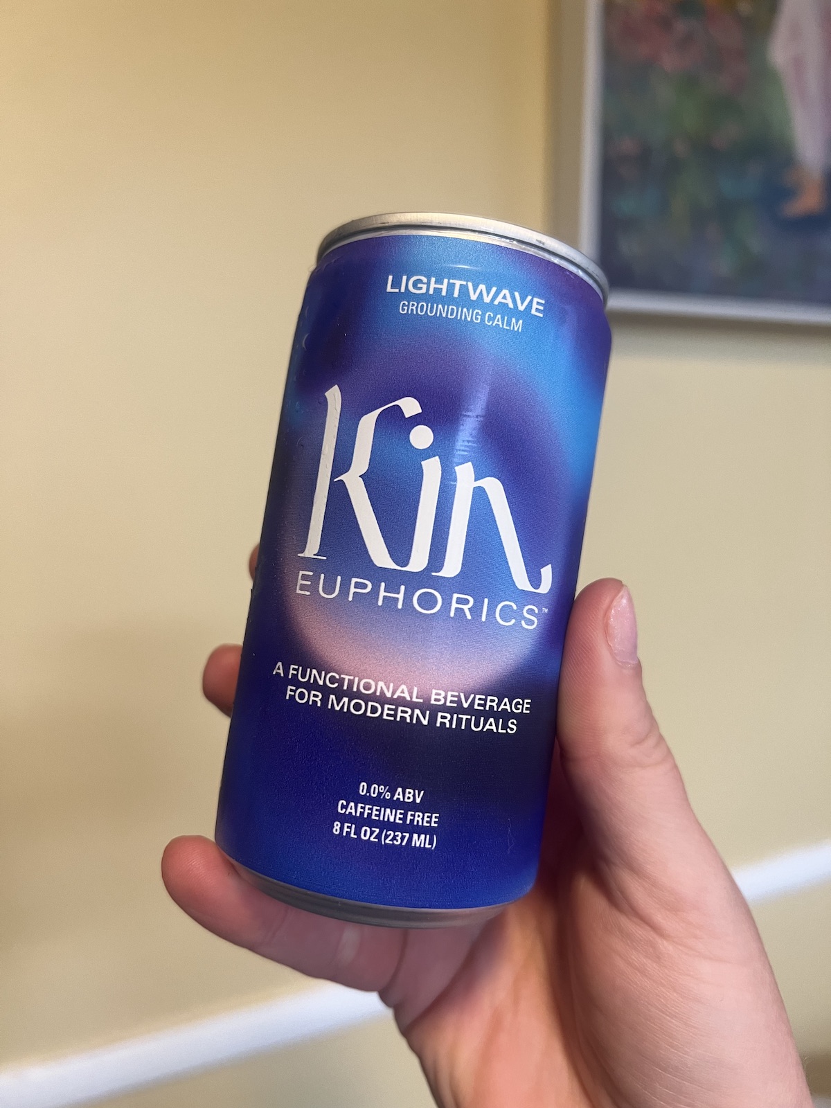 A hand holding a can of Kin Euphorics Lightwave, described as a "functional beverage for modern rituals" with 0.0% ABV and caffeine-free, containing 8 fl oz (237 ml). Experience the unique essence of this Kin drink today.