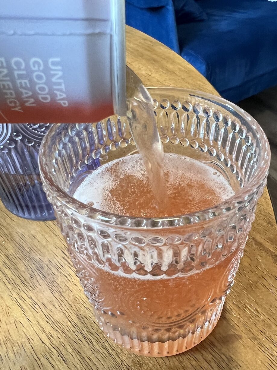 A canned beverage is being poured into a textured glass tumbler on a wooden surface. The drink is light pink in color. Another empty glass sits beside the tumbler.