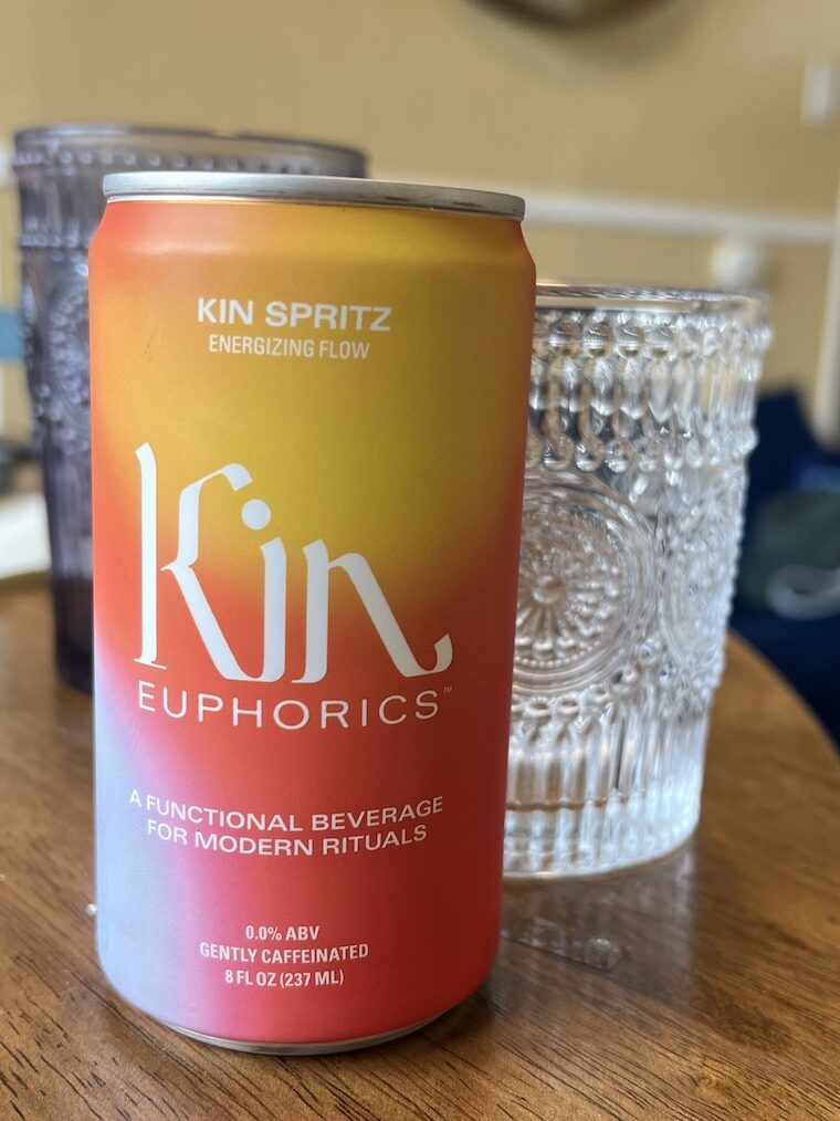 A can of Kin Euphorics Kin Spritz is placed next to an intricately patterned glass on a wooden surface. The beverage is labeled as a functional, gently caffeinated drink with 0.0% alcohol by volume.