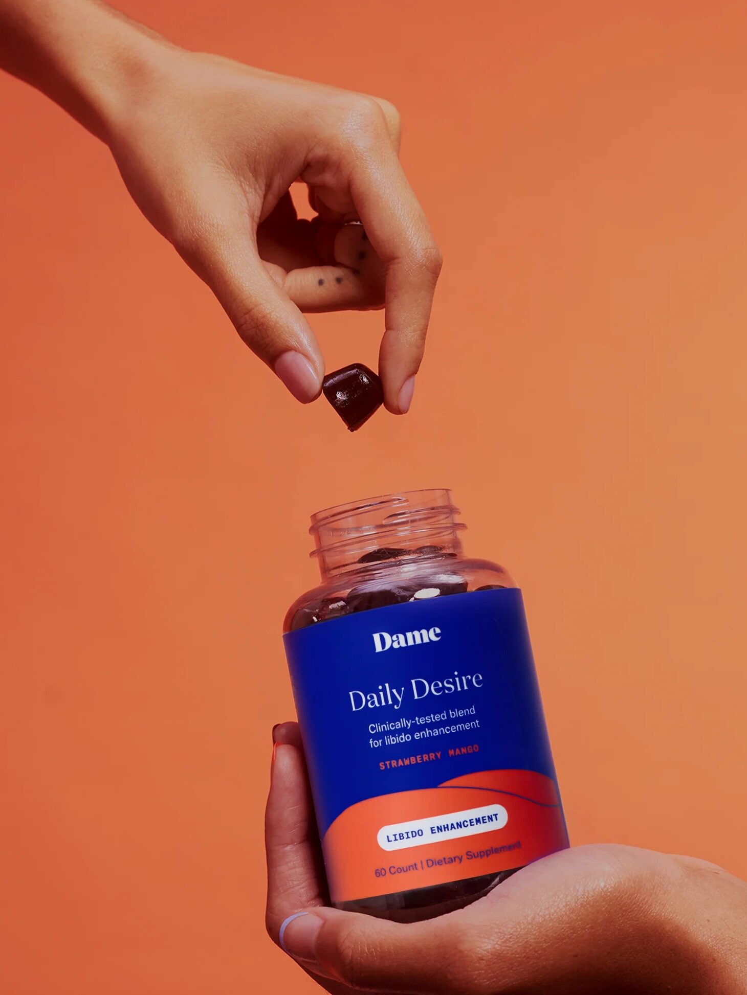 A hand is taking a gummy from a bottle labeled "Dame Daily Desire, Libido Enhancement." The bottle is being held by another hand against an orange background.