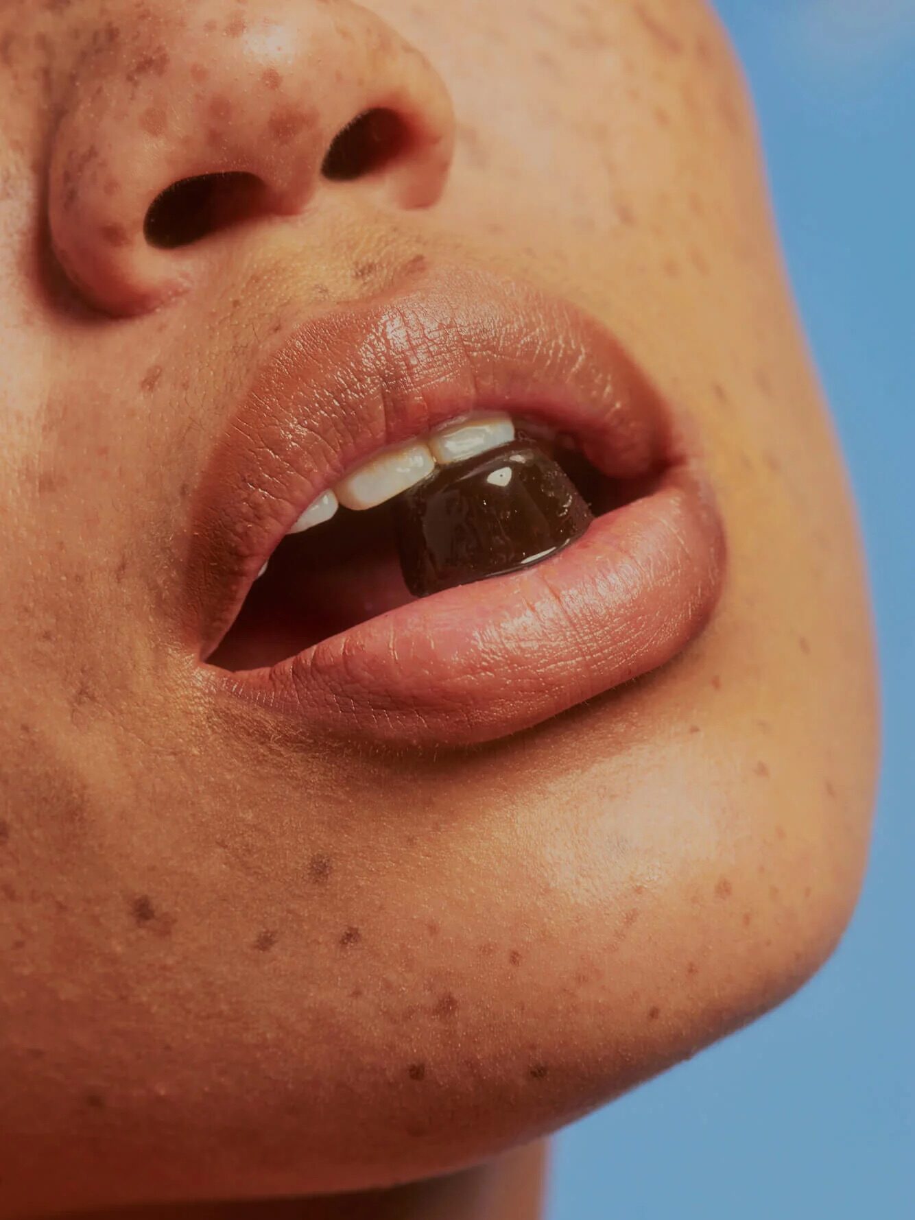 Close-up of a person's mouth slightly open, showing a piece of dark candy or gum between their teeth and lips. The skin around the mouth has freckles. The background is solid light blue.