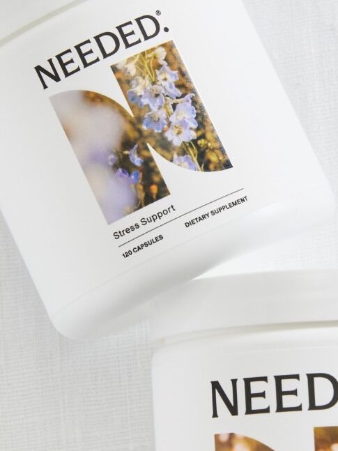 Two containers of dietary supplements labeled "NEEDED" are shown on a white surface with small white flowers around. The labels indicate "Stress Support" and "Sleep+Relaxation Support.