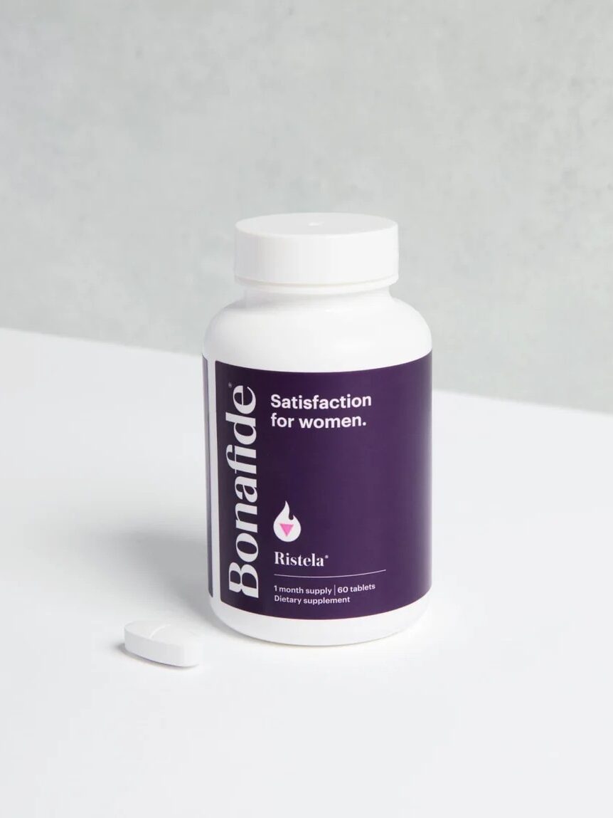 A bottle of Bonafide Ristela "Satisfaction for women" with one white pill next to it on a plain surface and a gray background.