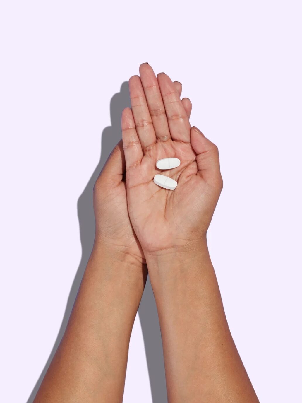 Two hands holding two white oblong pills against a light purple background.