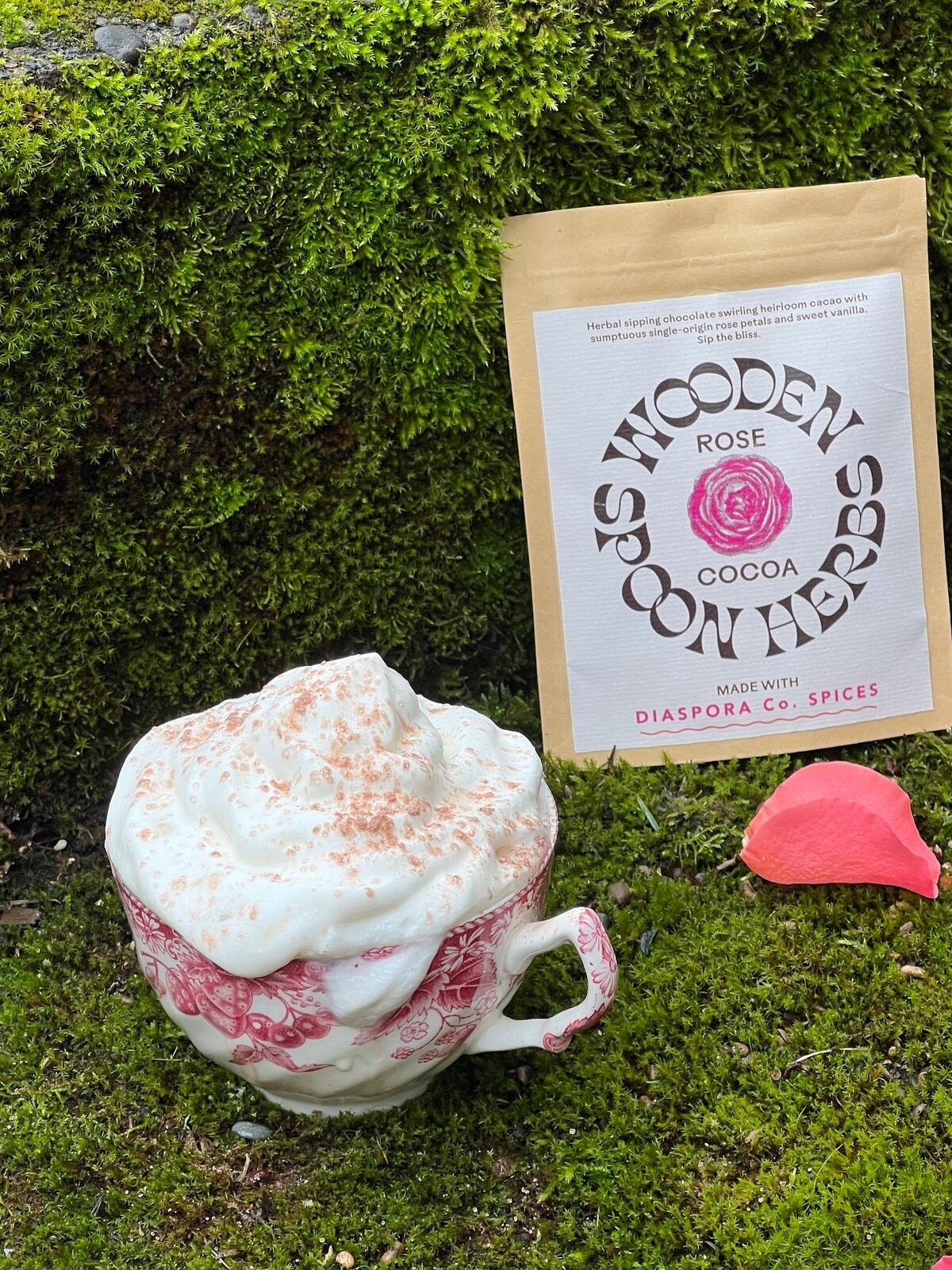 A patterned cup filled with a frothy beverage sits on mossy ground, accompanied by a packet labeled "Wooden Spoon Herbs Rose Cocoa." Rose petals are scattered around.