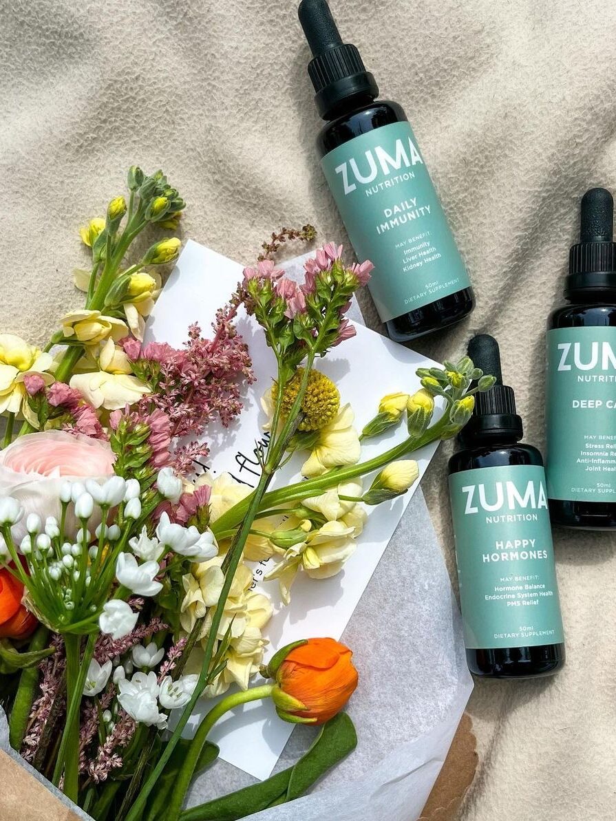 A bouquet of colorful flowers next to three green-labeled Zuma Nutrition bottles of Daily Immunity, Deep Calm, and Happy Hormones supplements. A partially visible card is beneath the flowers.