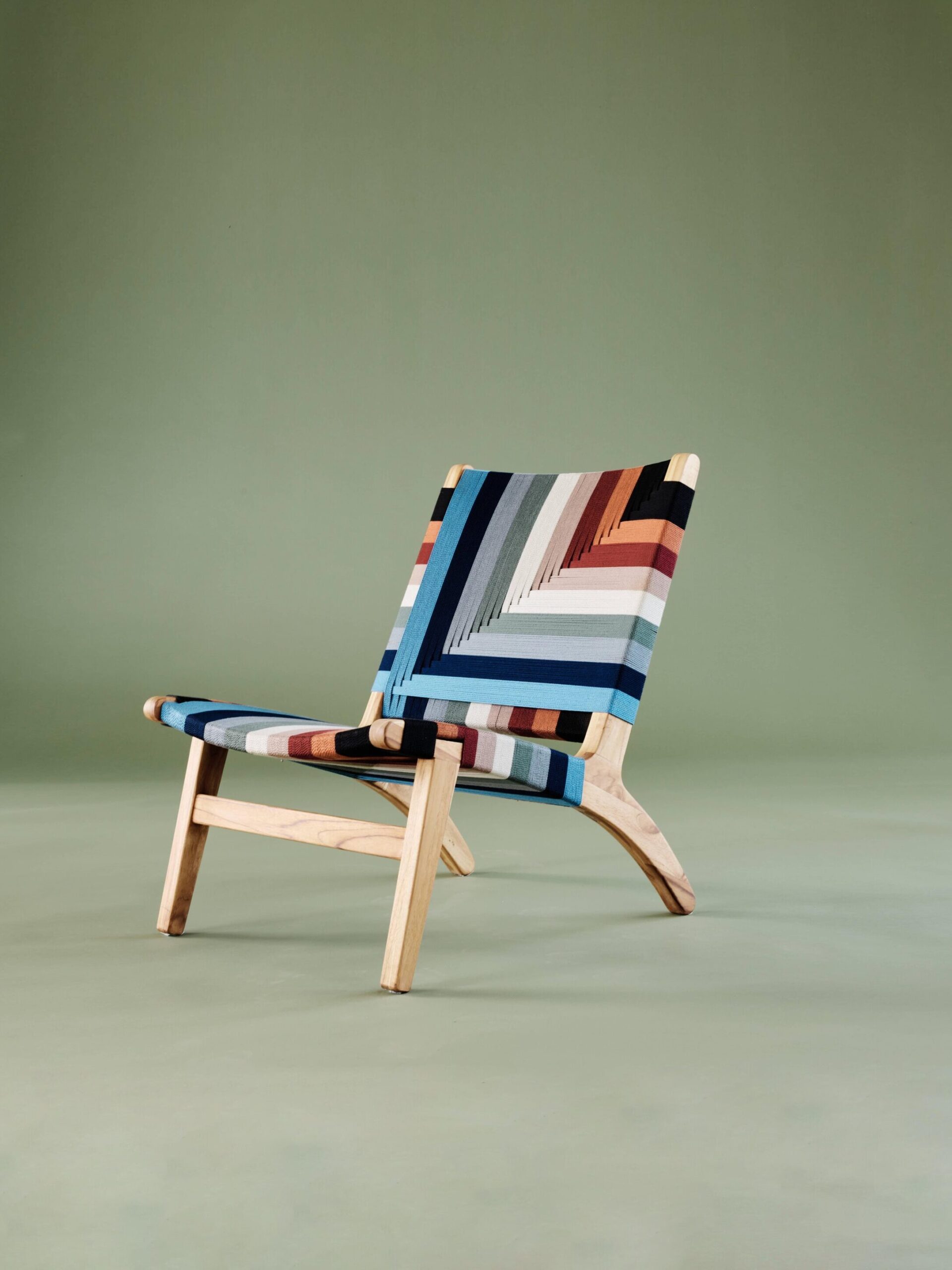 A wooden chair with a colorful, geometric patterned fabric backrest and seat, positioned against a muted green background.