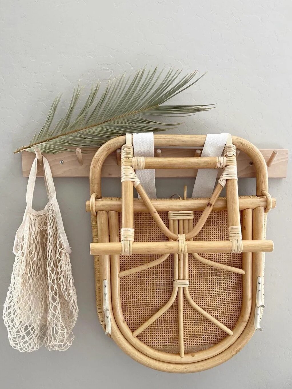 A wooden peg rack holds a folded rattan chair, an empty mesh bag, and a decorative palm leaf against a plain wall.
