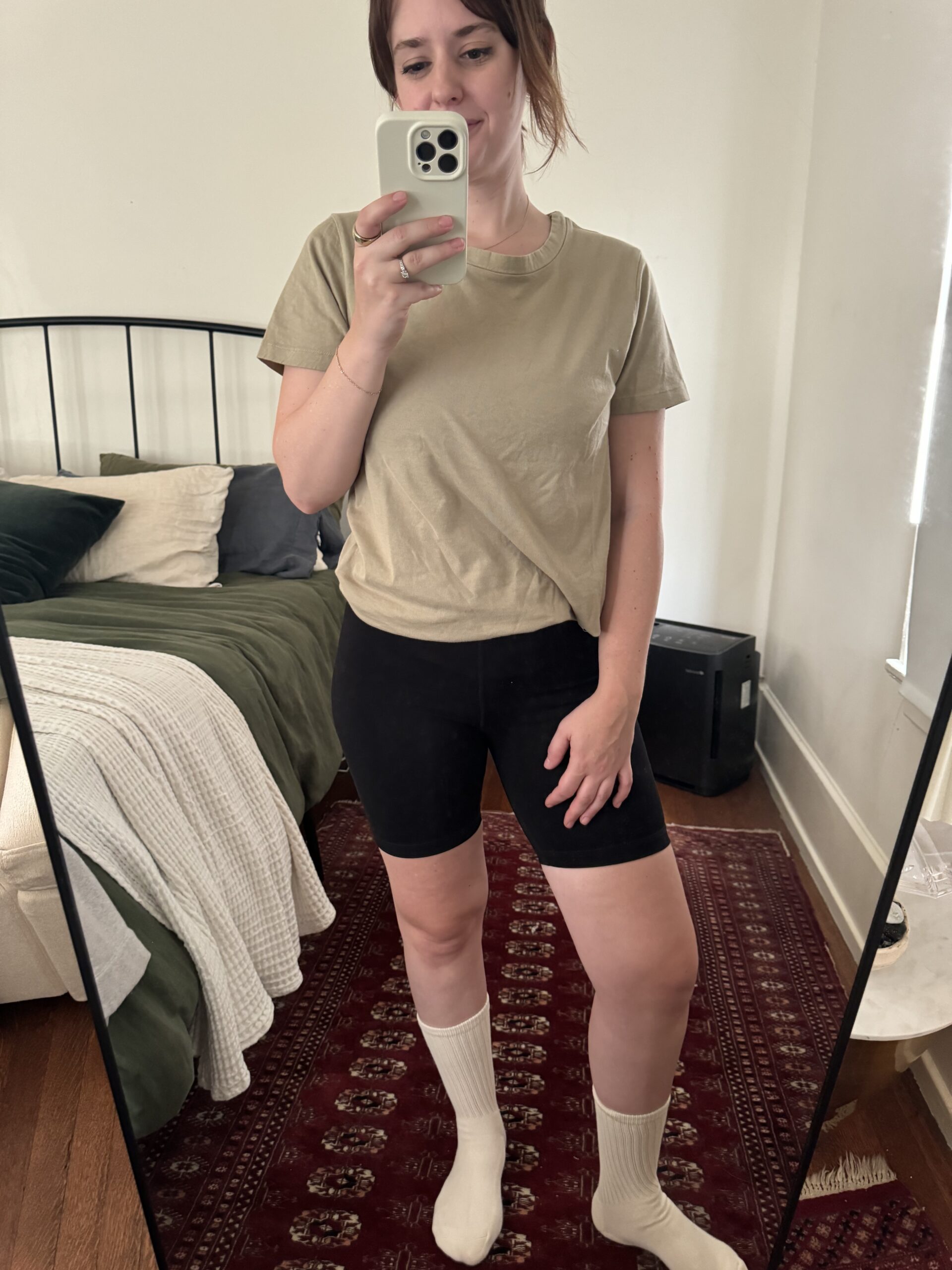 A person stands in front of a mirror taking a selfie. They are wearing a beige T-shirt, black shorts, and white socks. A bed with green and white bedding is visible in the background.