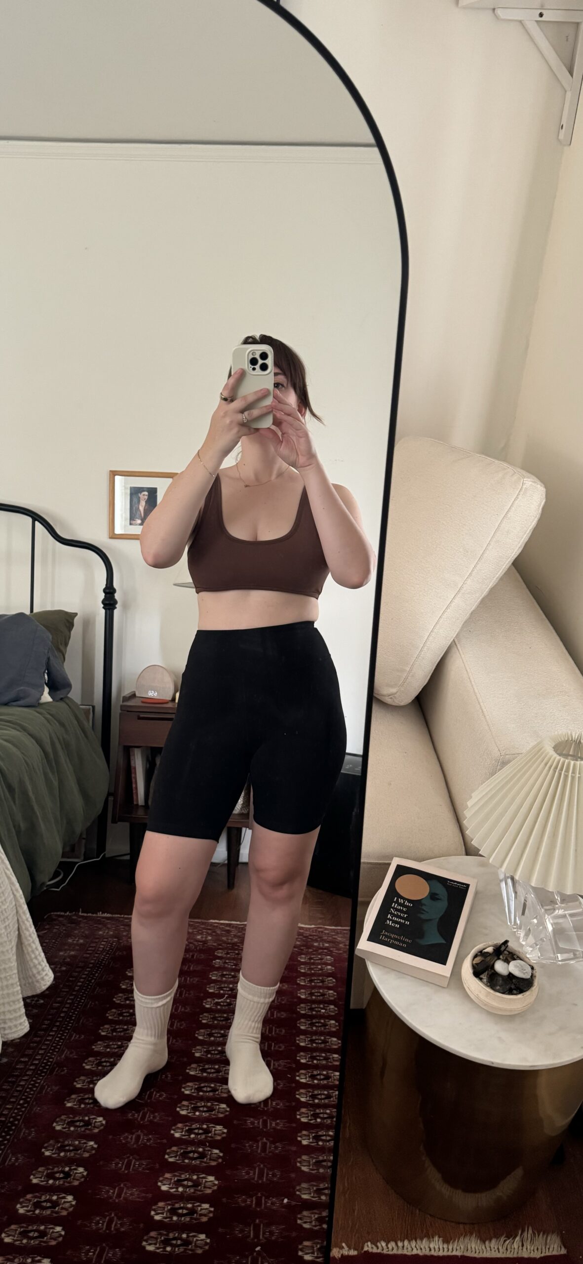 A person takes a mirror selfie in a bedroom, wearing a brown sports bra, black shorts, white socks, and holding a smartphone. The room has a bed, a green blanket, a beige sofa, and a side table with items.