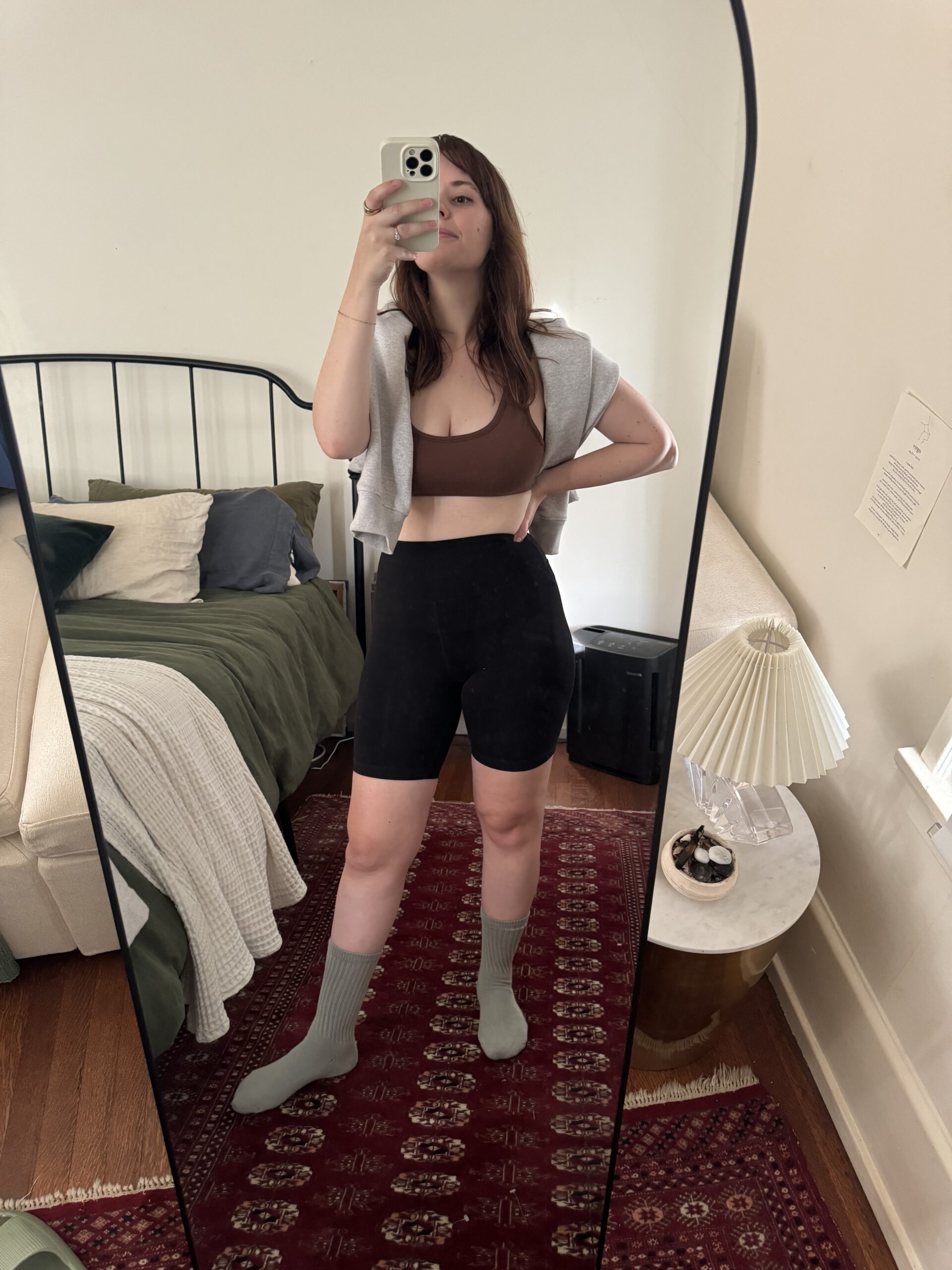 A woman takes a mirror selfie in a bedroom, wearing a sports bra, shorts, a hoodie, and socks. The bed is made with green and white bedding, and a bedside table with a lamp is in the background.