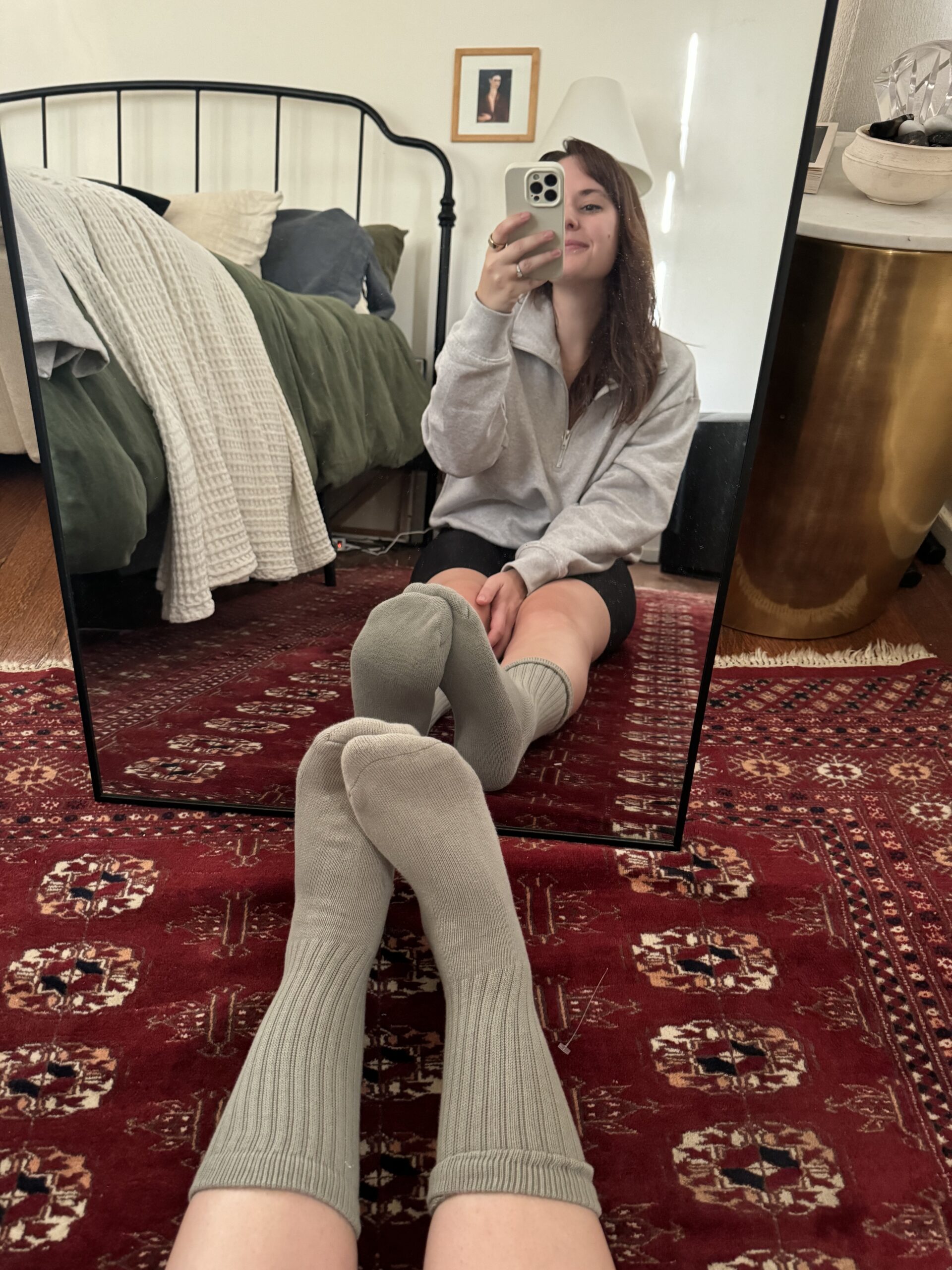 Person sitting on the floor in front of a mirror, taking a selfie. They are wearing a gray sweatshirt, dark shorts, and gray socks, with legs crossed. Bed and pillows in the background.