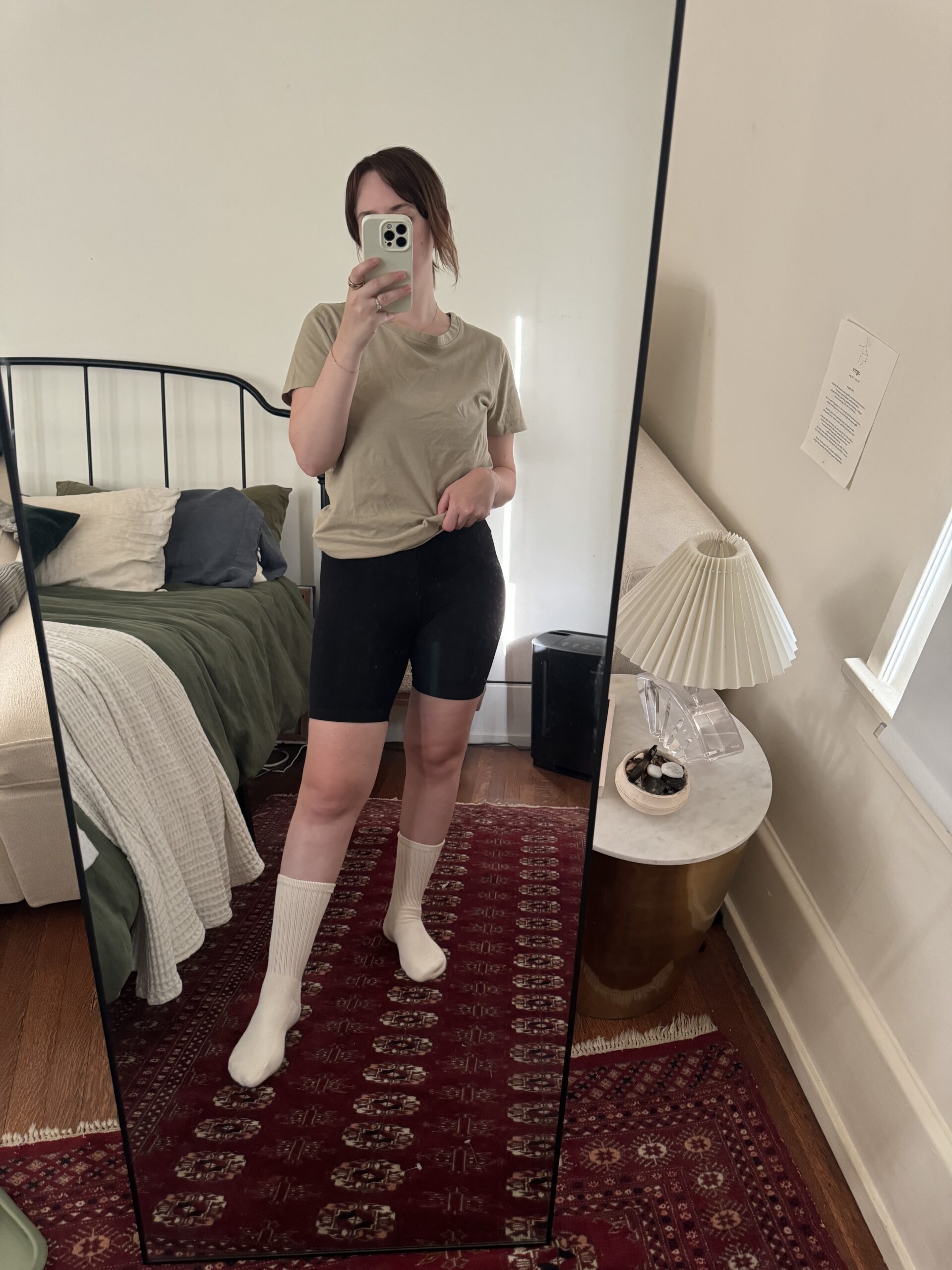 A person is taking a mirror selfie in a bedroom, wearing a beige t-shirt, black shorts, and white socks. The room features a bed with green and white bedding, a nightstand, and a lamp.