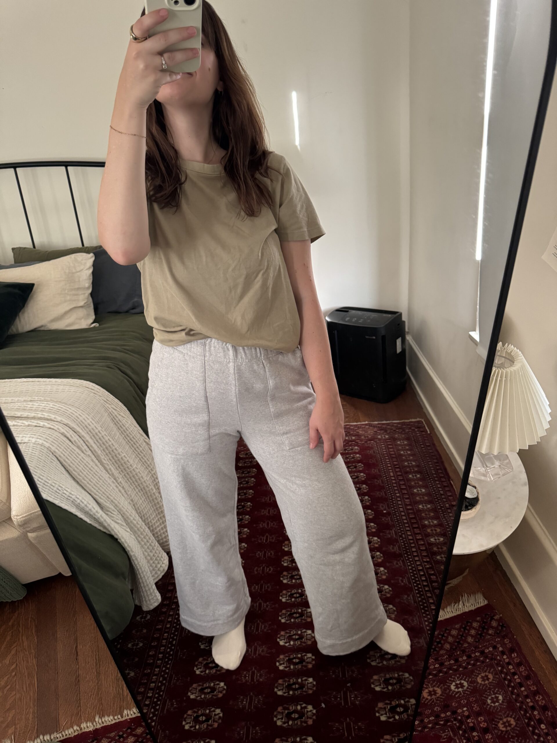 A person stands in front of a mirror taking a selfie, wearing a beige t-shirt and gray sweatpants. The room includes a bed with green and white bedding, a lamp on a round table, and a red rug.