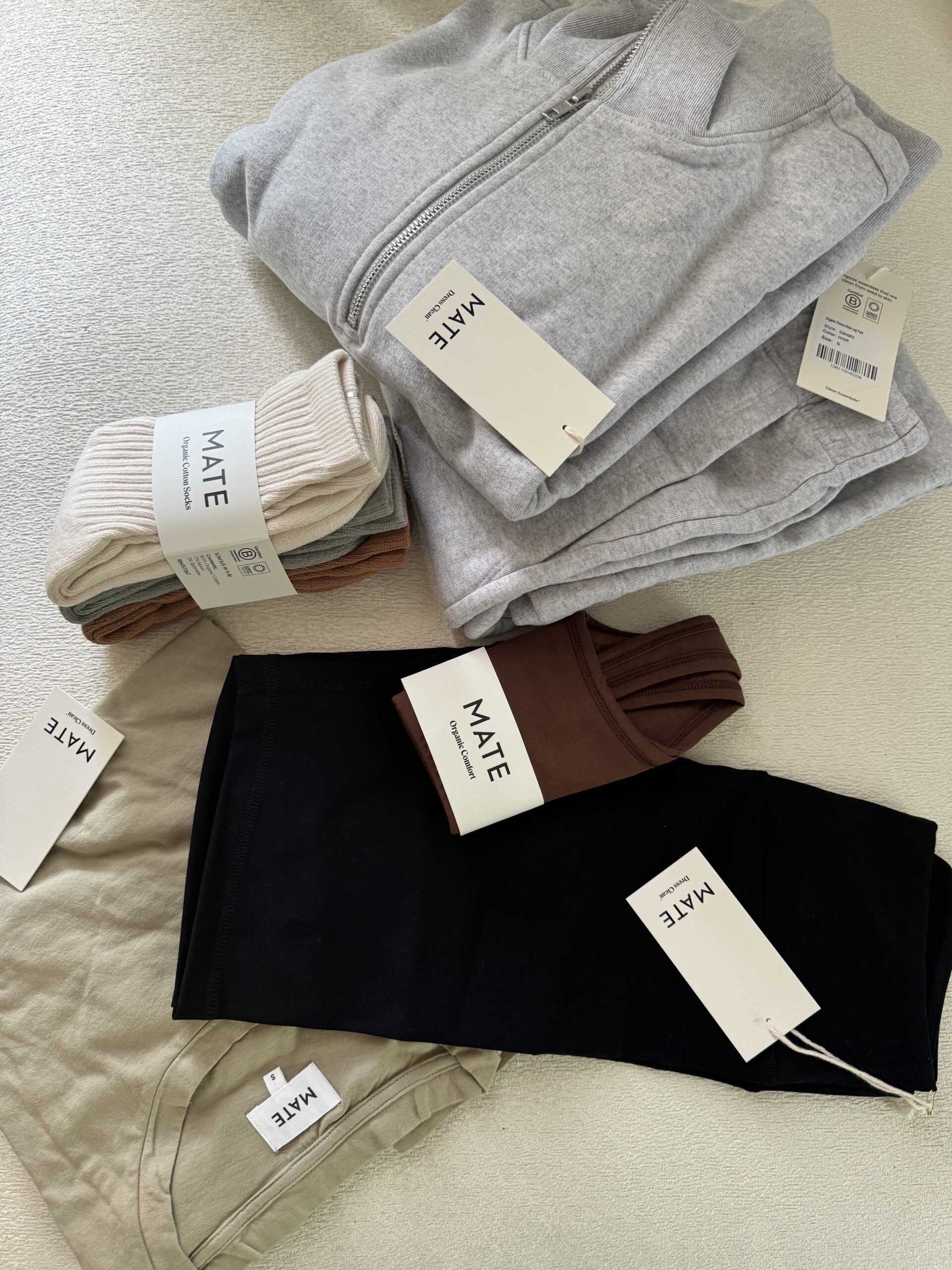 A collection of clothing items including a gray hoodie, beige and black pants, and several pairs of socks, all tagged with the brand "MATE".