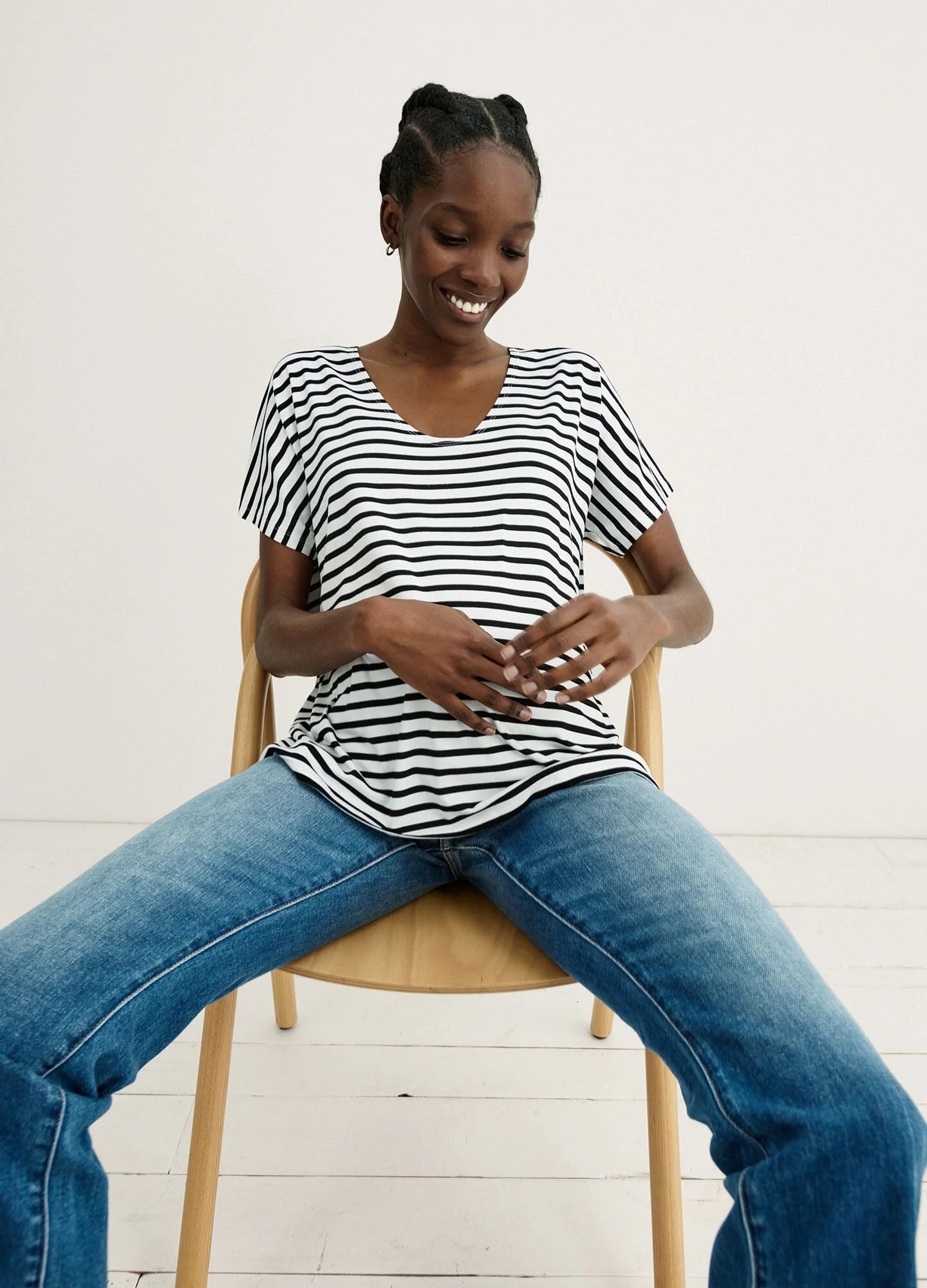 A person wearing a black and white striped shirt and blue jeans sits on a wooden chair, smiling with hands resting on their belly.